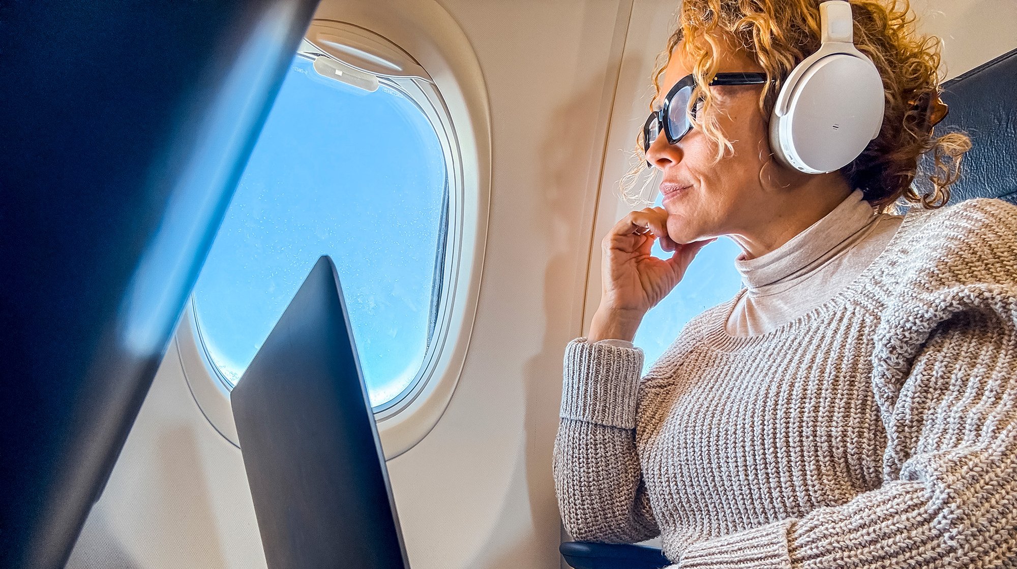 Person wearing headphones and sunglasses, looking out an airplane window, with a laptop on their lap