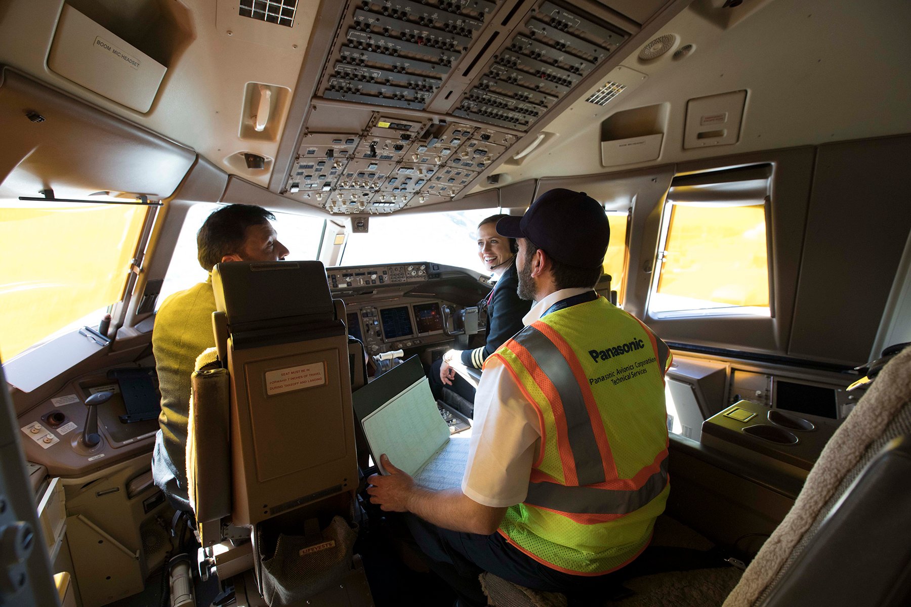 Three men in an airplane cockpit, two seated and one standing, surrounded by control panels and displays