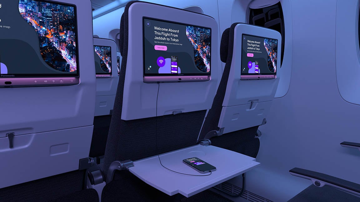 Airplane cabin with seats showing screens with a welcome message for Jeddah to Tokyo A smartphone on a tray table