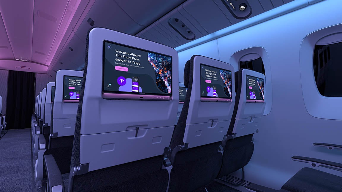 Cabin with seatback screens showing welcome message, under blue-purple lighting