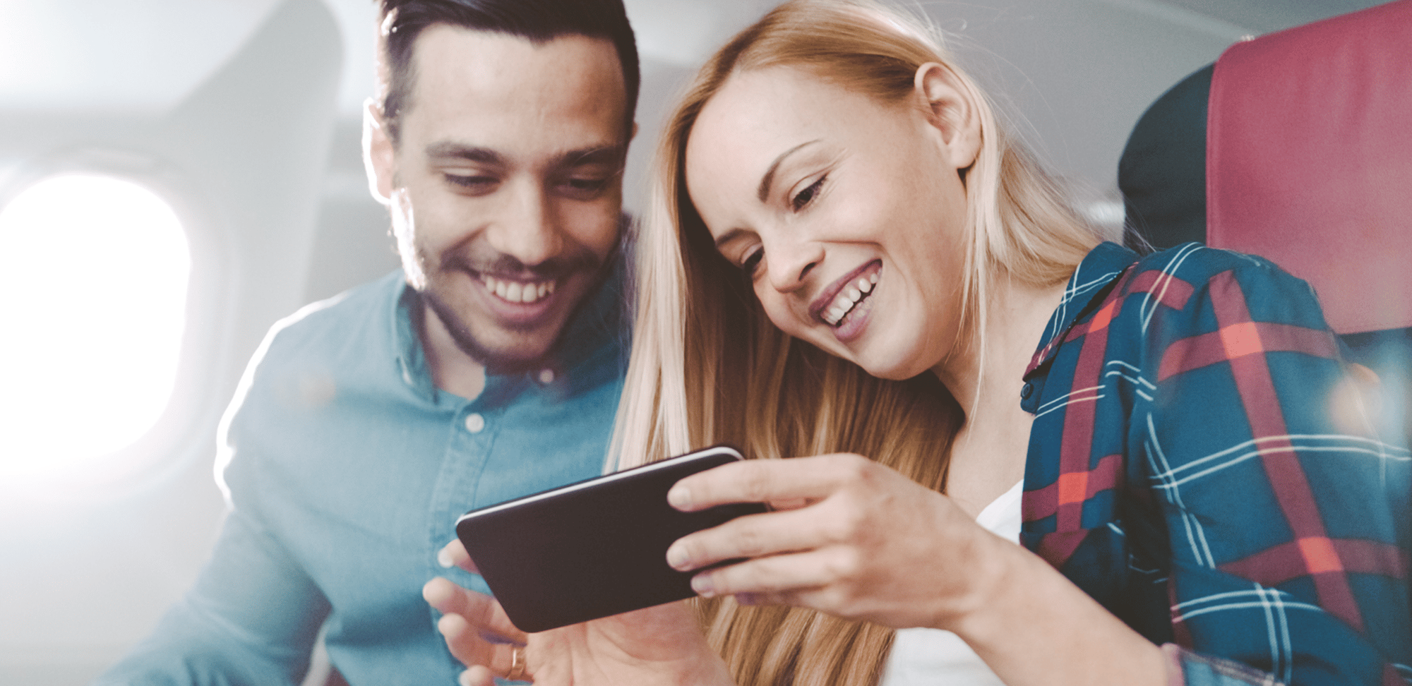 Two people on an airplane smiling and looking at a smartphone.