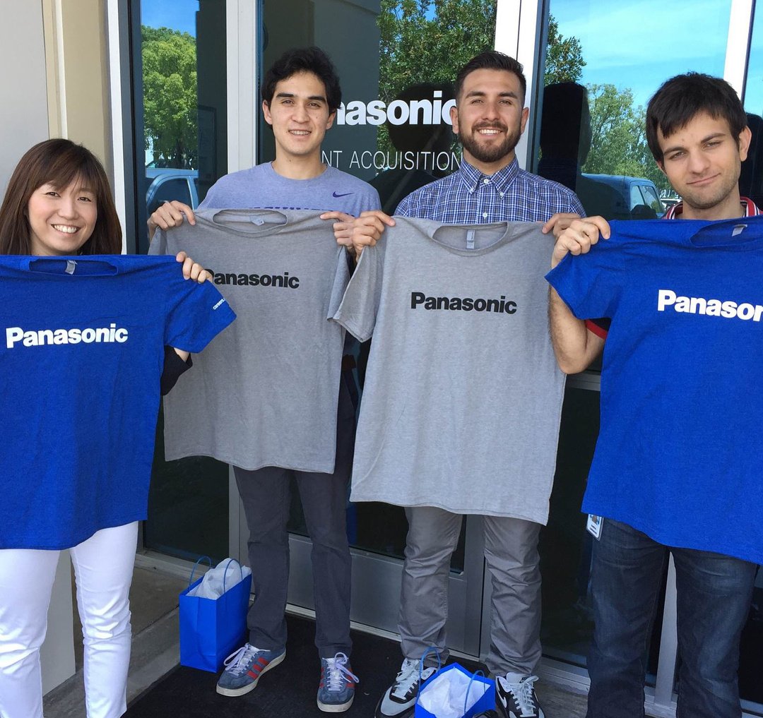 A group of four people stand holding Panasonic T-shirts in front of a glass door with the Panasonic logo