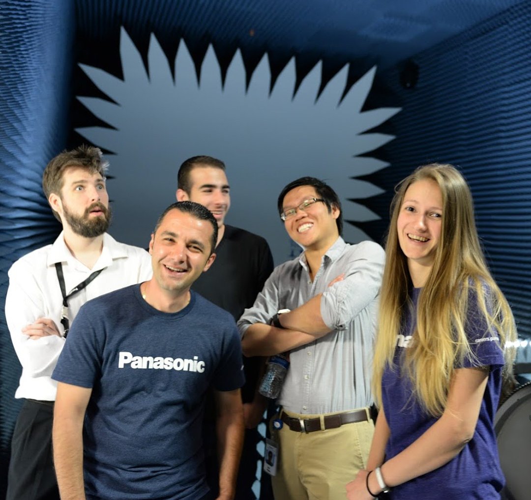 Group of five people smiling in front of a blue, soundproof background with "Panasonic" T-shirts visible