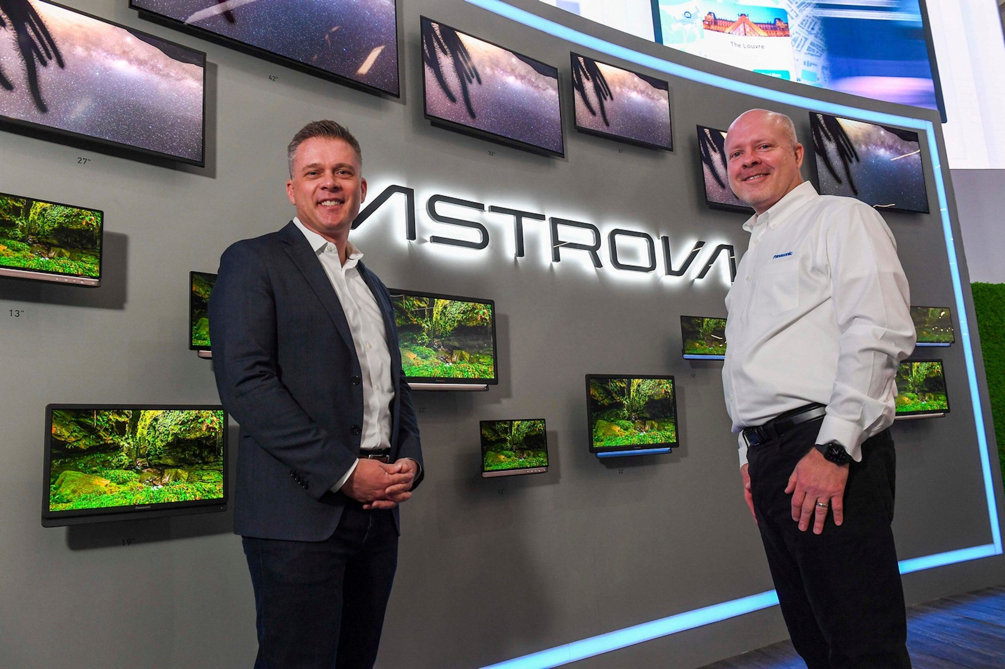 Two men smiling in front of a display wall showing starry sky and forest; illuminated "ASTROVA" sign